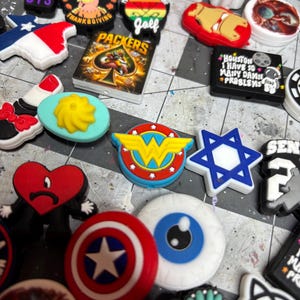 May include: A collection of colorful shoe charms with various designs. Notable charms include a Wonder Woman logo, a Captain America shield, a Star of David, and charms with text like "BTS" and "Packers".