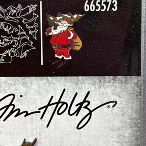 May include: A black surface featuring the number "665573" in white and a cartoon Santa Claus in a red suit with a gold sack. The image also includes the signature "Tim Holtz".