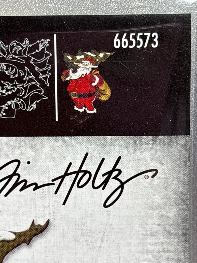 May include: A black surface featuring the number "665573" in white and a cartoon Santa Claus in a red suit with a gold sack. The image also includes the signature "Tim Holtz".