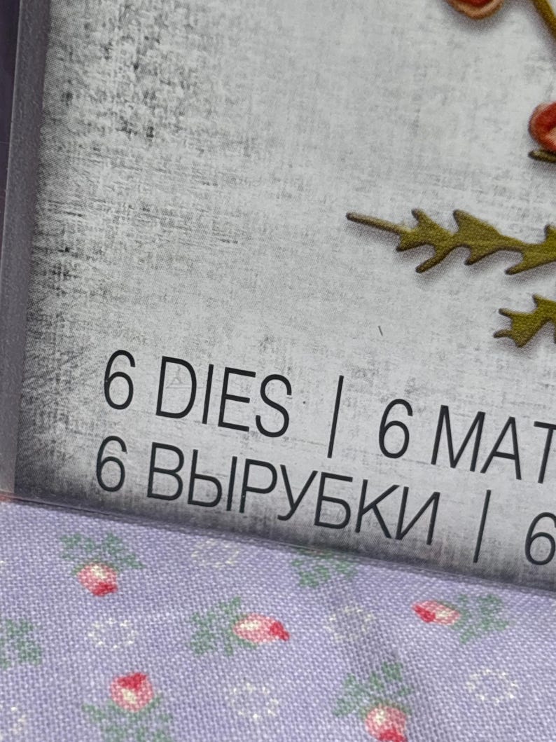 May include: Close-up of a package with the text "6 DIES | 6 MAT". The background is a light grey with a floral pattern. The text is in black. The package is slightly angled.