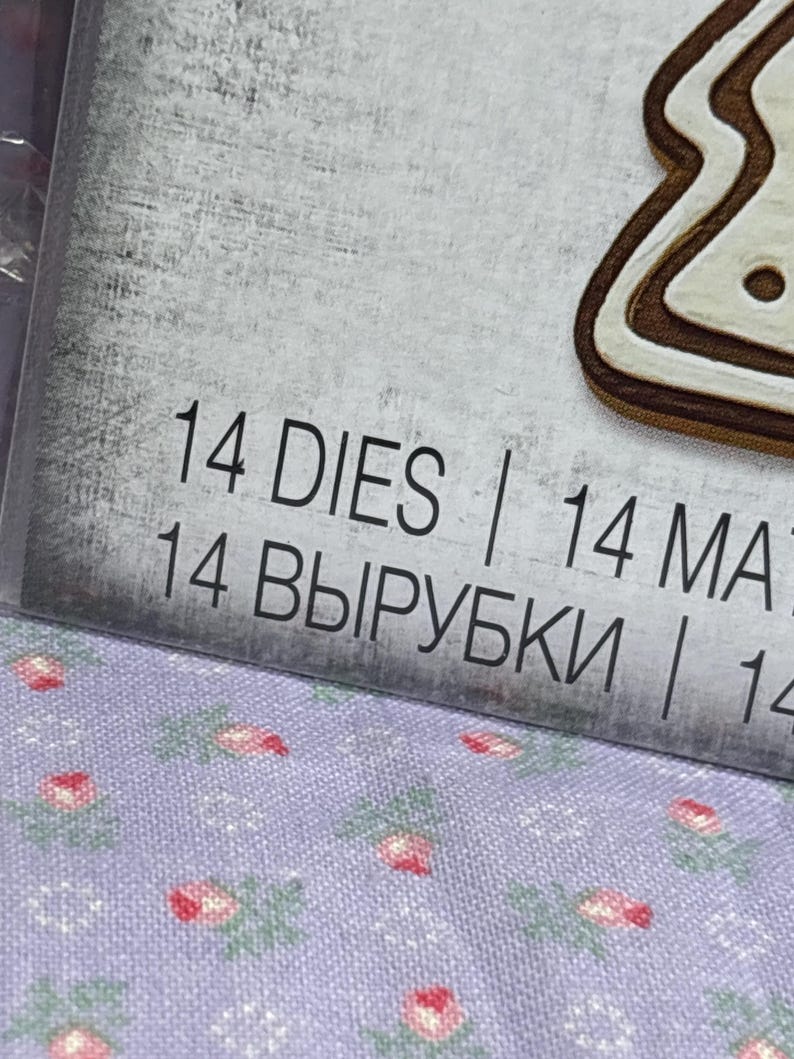 Puede incluir: Primer plano de un paquete con el texto "14 DIES" y "14 MAT". El fondo es una superficie gris claro texturizada. Se ve un troquel con forma de galleta de jengibre. La parte inferior de la imagen muestra una tela con estampado floral.