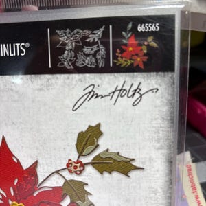 May include: A clear plastic package containing a die-cut set with a poinsettia and holly design. The package features the brand name "Sizzix" and the signature "Jim Holtz". The poinsettia is red with yellow accents, and the holly is green.