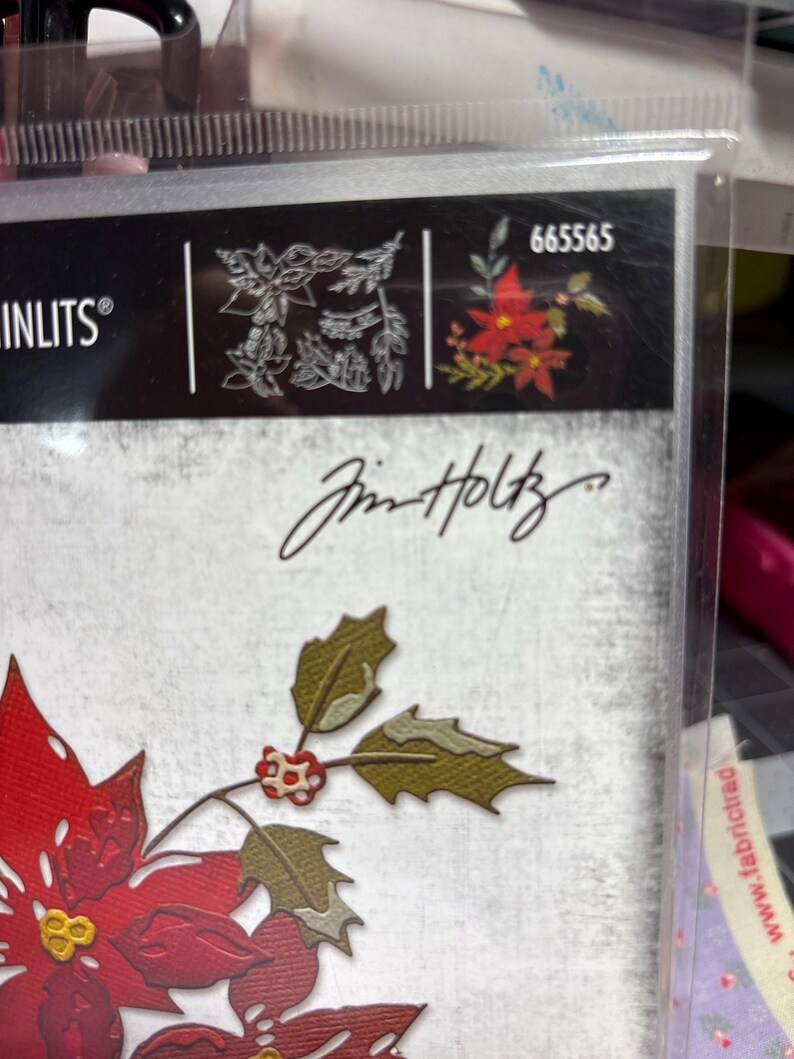 May include: A clear plastic package containing a die-cut set with a poinsettia and holly design. The package features the brand name "Sizzix" and the signature "Jim Holtz". The poinsettia is red with yellow accents, and the holly is green.