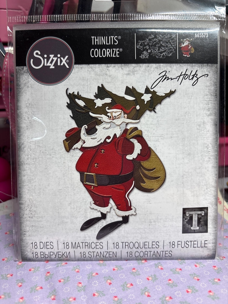 May include: A Sizzix Thinlits Colorize die set with a Santa Claus design carrying a Christmas tree. The Santa Claus is in a red suit with a black belt and boots. The package includes 18 dies and the signature of Jim Holtz.