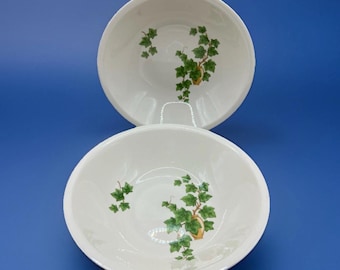 May include: Two white ceramic bowls with a green ivy design. The bowls have a rounded shape and a slightly flared rim. The ivy design features green leaves and a small, gold-colored pot.