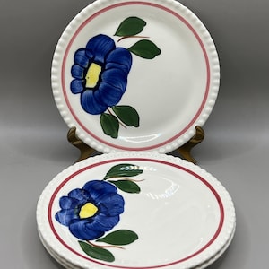 May include: Set of four white ceramic plates with a blue flower and green leaves design. The plates have a red and white scalloped edge.
