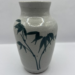 May include: A speckled, off-white ceramic vase with a slightly flared rim. Dark green bamboo leaves and stalks are painted on the vase. The vase has a rounded body and a narrow neck, suitable for displaying flowers or as a decorative piece.