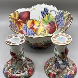 May include: A decorative ceramic fruit bowl and two matching candle holders. The bowl and candle holders feature a hand-painted design of various fruits, including grapes, pears, and apples, with a gold trim. The color palette includes red, green, yellow, and purple.