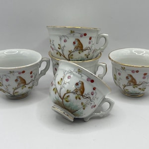May include: A collection of six white porcelain teacups. Each cup features a gold rim and a painted design of a bird perched on a branch with red berries. The cups have small handles.