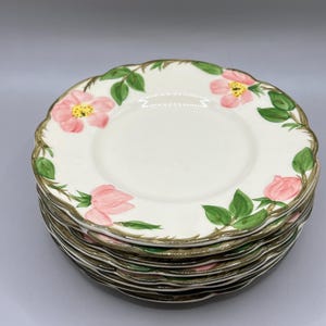 May include: A stack of white ceramic plates with a scalloped edge. Each plate is decorated with pink rose blossoms, green leaves, and a brown rim. The plates are stacked on a light gray surface.