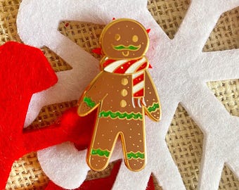 May include: A gingerbread man enamel pin with a gold outline, wearing a red and white striped scarf and a green mustache. The pin is set against a white snowflake and red felt background.