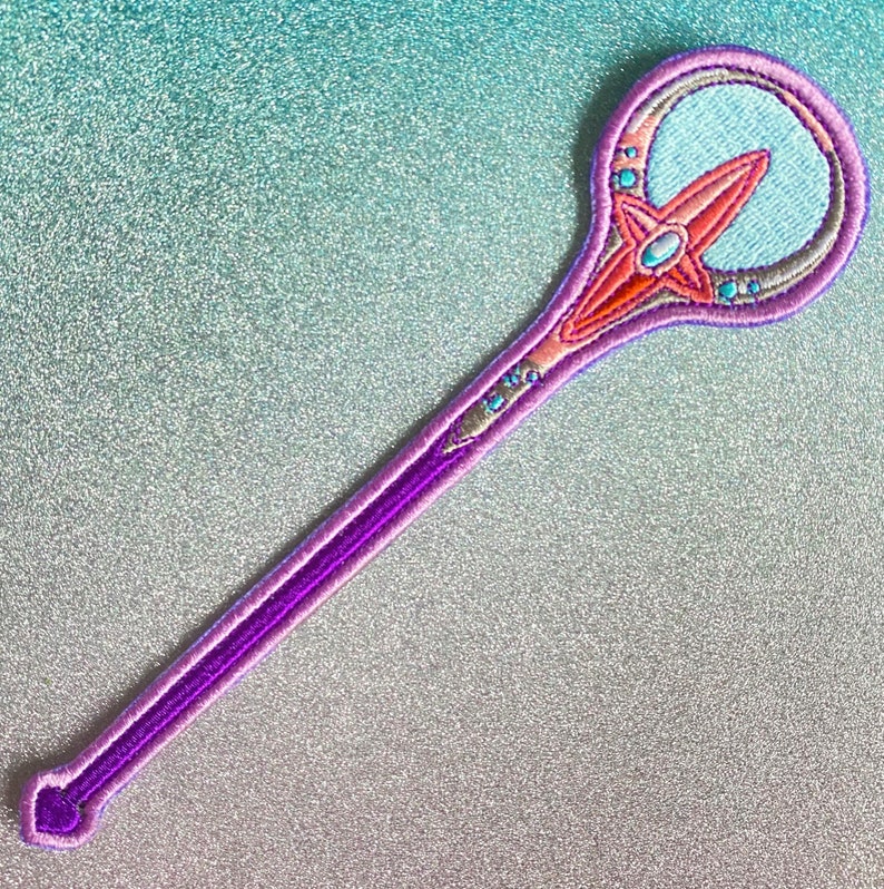 May include: A purple and pink embroidered wand with a blue and orange star design. The wand has a rounded tip and a long, thin handle.