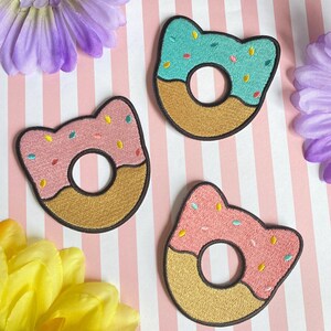 Puede incluir: Tres parches bordados para planchar con forma de rosquillas con glaseado rosa, azul y marrón y espolvoreados de colores.