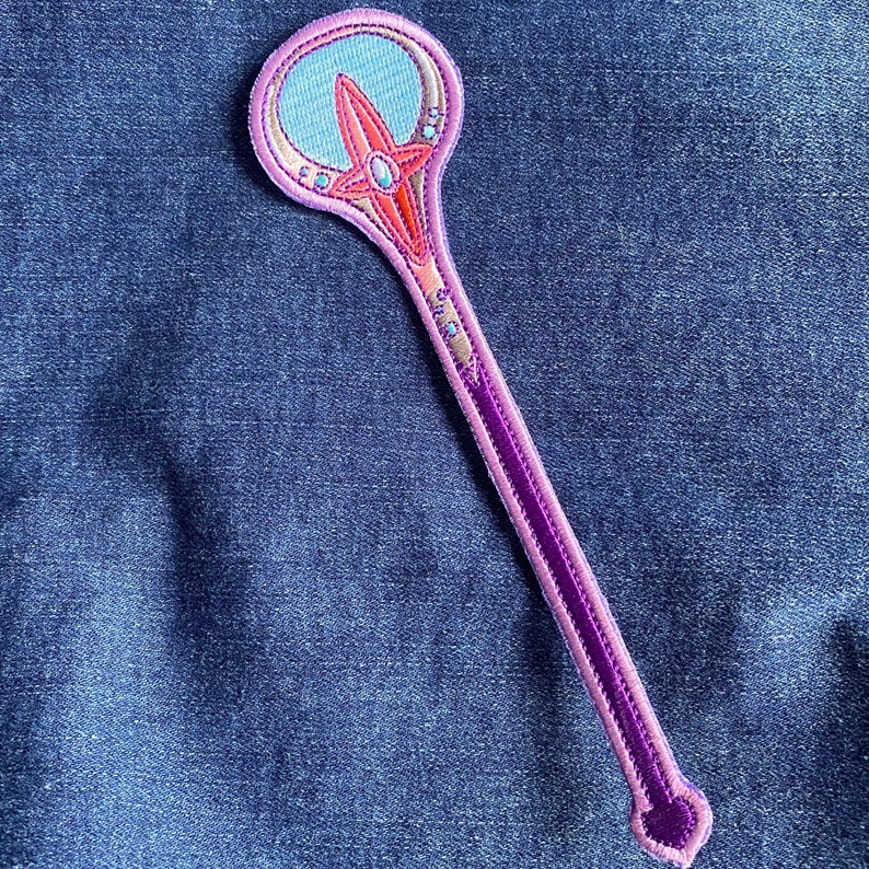 May include: A purple and pink embroidered wand with a blue and pink star design. The wand has a round top with a blue and pink star design.