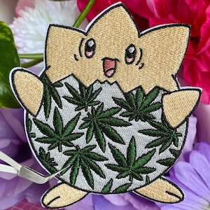 May include: Embroidered patch featuring a cartoon character emerging from an egg. The eggshell is tan, and the character has a smiling face. The egg's interior is white with green cannabis leaves.