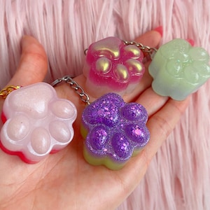 May include: Four colourful paw print keychains. The keychains are in the shape of a paw print and are in pink, purple, and green. The keychains have a glittery or iridescent finish and are attached to silver keychains.