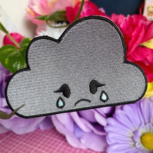 May include: A grey embroidered patch shaped like a cloud with a sad face and two teardrops. The cloud has black eyes and a black frown.
