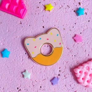 Könnte beinhalten: Eine rosa und orangefarbene Emaille-Anstecknadel in Form eines Donuts mit Streuseln. Die Anstecknadel befindet sich auf einem rosa Hintergrund mit kleinen sternförmigen Verzierungen.