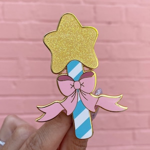 Puede incluir: Un pin de tono dorado con una estrella amarilla brillante en la parte superior de un bastón de caramelo azul y blanco a rayas con un lazo rosa.