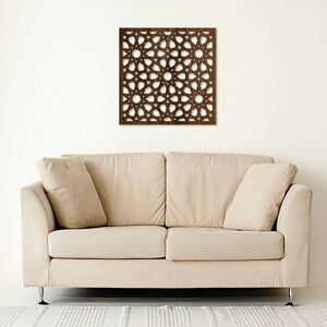 K&ouml;nnte beinhalten: Eine braune Holz-Wanddekoration mit geometrischem Muster. Die Dekoration h&auml;ngt an einer wei&szlig;en Wand &uuml;ber einem beigen Sofa.