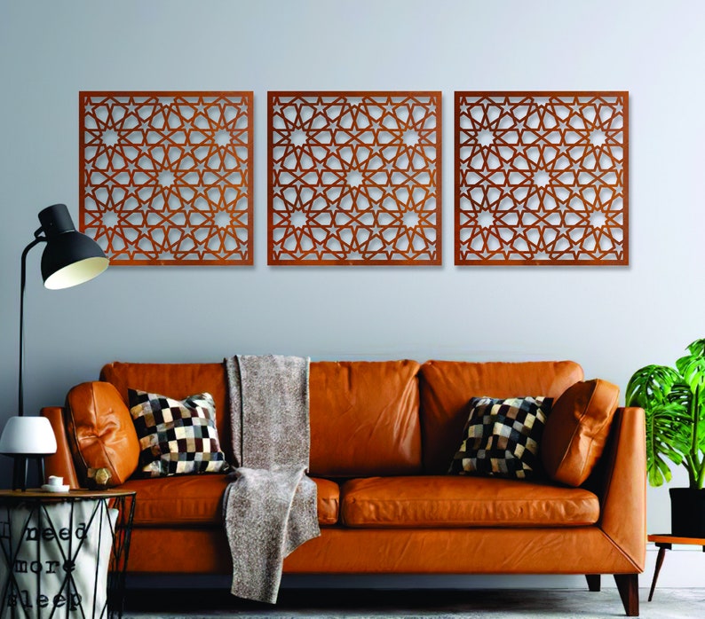 K&ouml;nnte beinhalten: Drei braune Holz-Wanddekorationspaneele mit einem geometrischen Sternmuster. Die Paneele h&auml;ngen an einer hellgrauen Wand &uuml;ber einem braunen Ledersofa.