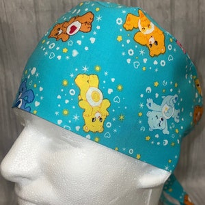 May include: A turquoise blue surgical scrub hat with a Care Bears print. The hat features a variety of Care Bears characters, including Cheer Bear, Funshine Bear, and Grumpy Bear. The hat has a tie at the back.