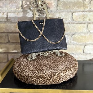 Op de afbeelding: Zwarte clutch met een gouden ketting, tentoongesteld op een decoratieve standaard. De clutch heeft een textuur met slangenpatroon. De standaard staat op een kussen met luipaardprint.