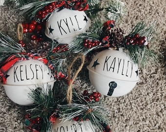 May include: Four white ceramic Christmas bells with black star and bell details. Each bell is decorated with a black name: Kourt, Kelsey, Kayley, and Kyle. The bells are surrounded by green pine branches with red berries and plaid ribbon.