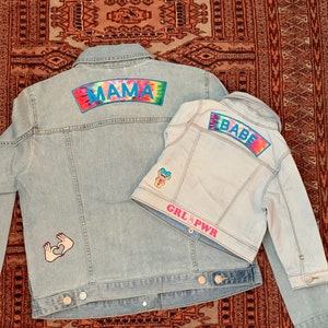 May include: Two denim jackets with colourful patches. The larger jacket has a "MAMA" patch and a pink heart hand gesture patch. The smaller jacket has a "BABE" patch and a "GIRL POWER" patch.
