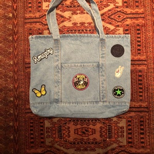 May include: A blue denim tote bag with various patches, including a butterfly, a peace sign, a black circle with a white symbol, and a pink and white patch that says "Good Child". The bag also has a white patch that says "Amazing" on the front.