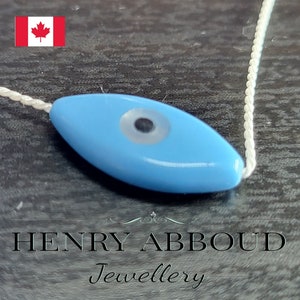 May include: A pale blue, eye-shaped bead with a white and black eye design, strung on a white cord. The bead is the focal point, with the text "HENRY ABBOUD Jewellery" below. The Canadian flag is in the upper left corner.