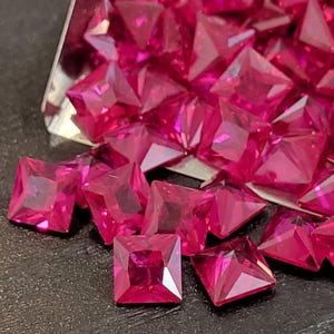 May include: Close-up of a collection of bright pink square-cut gemstones. The gems are faceted, reflecting light, and appear to be loose. The gems are scattered on a dark surface, with some in a small metal tray.