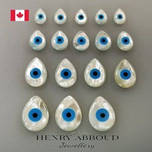 May include: A collection of 18 white mother of pearl evil eye cabochons with blue and black accents. The cabochons are in the shape of a teardrop.