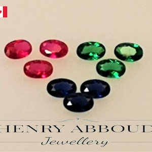 Puede incluir: Surtido de piedras preciosas ovaladas en rojo, verde y azul. Las piedras preciosas están dispuestas en un patrón sobre una superficie blanca. Henry Abboud Joyería.