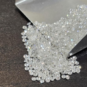 May include: A pile of small, clear diamonds cascading from a silver scoop onto a dark surface. The diamonds glisten, reflecting light. The image provides a close-up view of the gemstones, emphasising their clarity and sparkle.