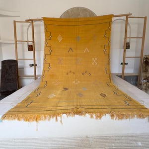 Pu&ograve; includere: Tappeto tessuto giallo senape con motivi geometrici e frange. Il tappeto presenta disegni a rombi, croci e frecce in vari colori. Il tappeto &egrave; esposto su una superficie bianca, con una cornice in legno sullo sfondo.
