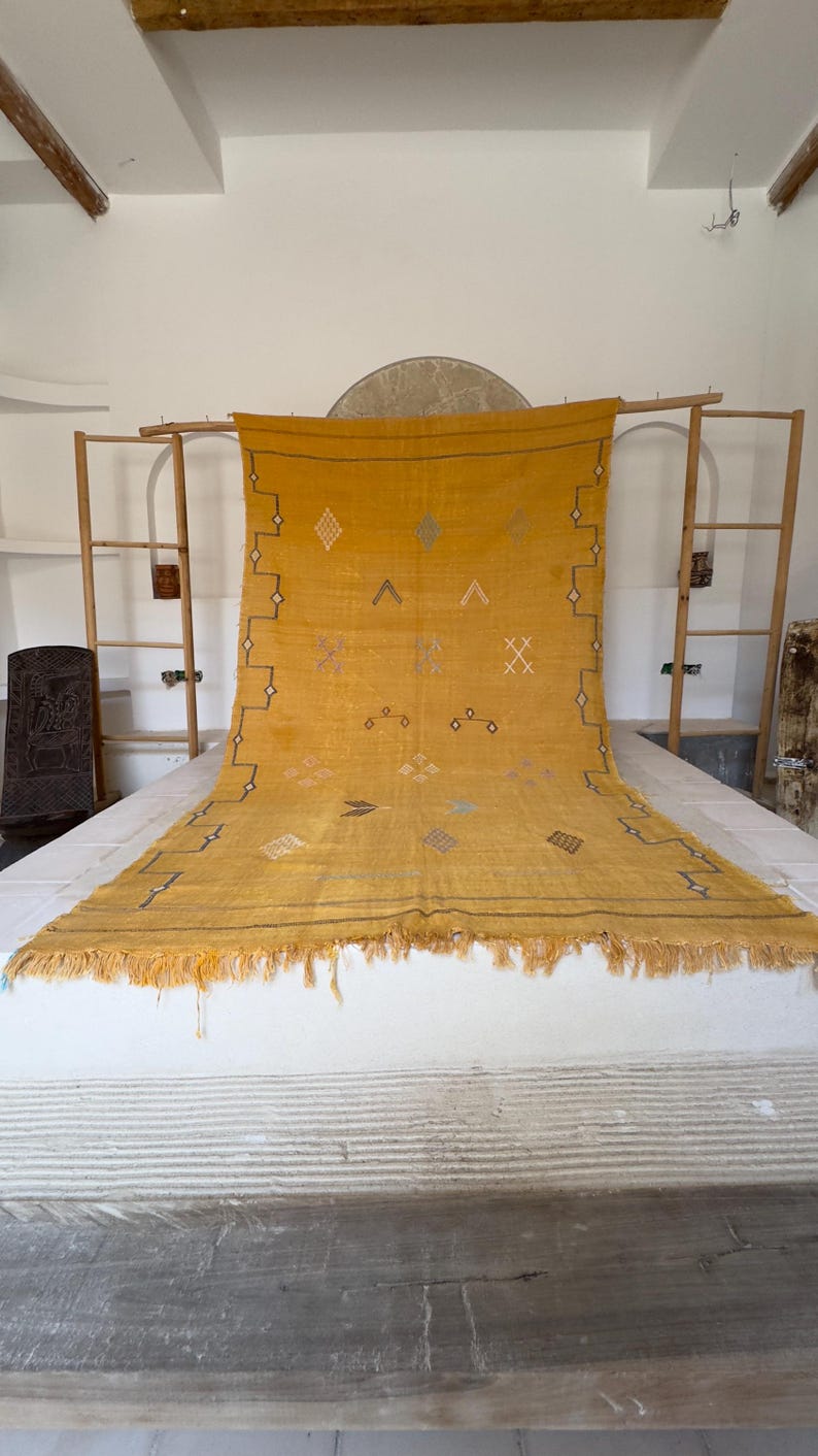 Pu&ograve; includere: Tappeto tessuto giallo senape con motivi geometrici e frange. Il tappeto presenta disegni a rombi, croci e frecce in vari colori. Il tappeto &egrave; esposto su una superficie bianca, con una cornice in legno sullo sfondo.