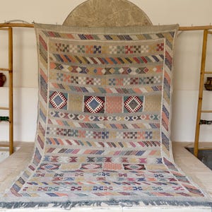 May include: A large, rectangular, beige rug with a geometric pattern in various colors, including blue, red, orange, and pink. The rug has a fringe along the bottom edge and is displayed on a wooden frame.