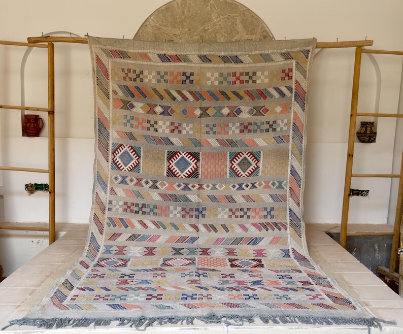 May include: A large, rectangular, beige rug with a geometric pattern in various colors, including blue, red, orange, and pink. The rug has a fringe along the bottom edge and is displayed on a wooden frame.
