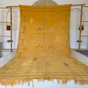 Pu&ograve; includere: Tappeto giallo senape con motivi geometrici e frange. Il tappeto &egrave; esposto su una superficie bianca, con una cornice in legno e una finestra sullo sfondo. Il tappeto presenta una serie di motivi a diamante, triangolo e croce.
