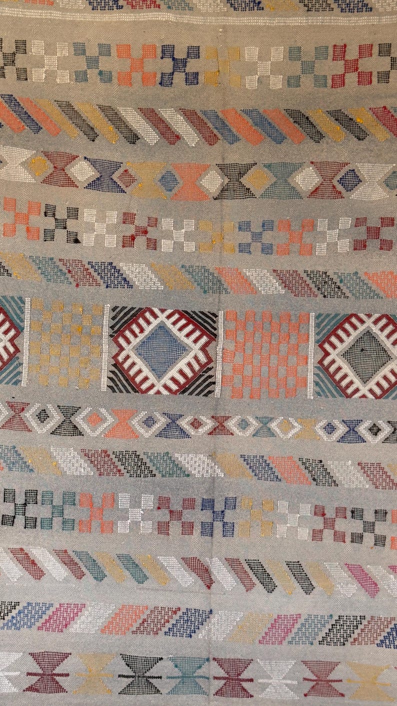 May include: Close-up of a woven textile with a light gray background. The design features repeating geometric patterns in various colors, including red, blue, orange, and yellow. The patterns include diamonds, squares, and diagonal stripes.