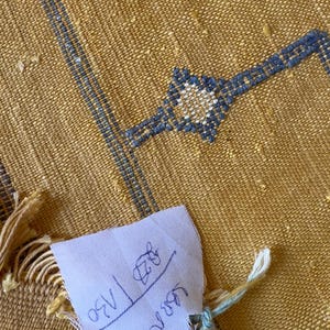 Pu&ograve; includere: Tessuto giallo senape tessuto con ricami geometrici blu. Una piccola etichetta bianca &egrave; attaccata con testo scritto a mano. Il tessuto ha un aspetto strutturato e un bordo sfrangiato. Il ricamo include un disegno a forma di stella.