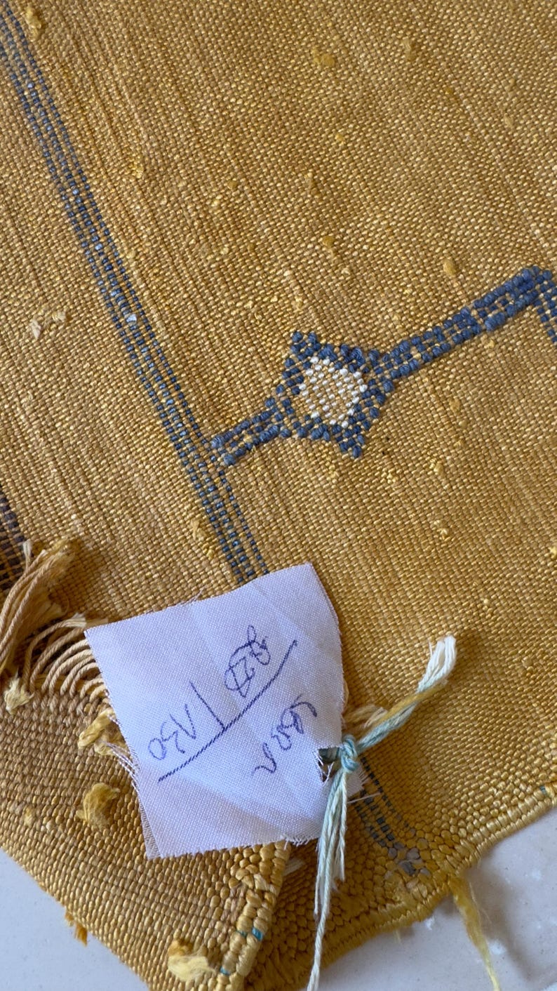 Pu&ograve; includere: Tessuto giallo senape tessuto con ricami geometrici blu. Una piccola etichetta bianca &egrave; attaccata con testo scritto a mano. Il tessuto ha un aspetto strutturato e un bordo sfrangiato. Il ricamo include un disegno a forma di stella.