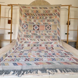 May include: A large, rectangular rug with a beige background and colorful geometric patterns. The design includes squares, diamonds, and stripes in shades of blue, pink, orange, and green. The rug has a fringed edge.