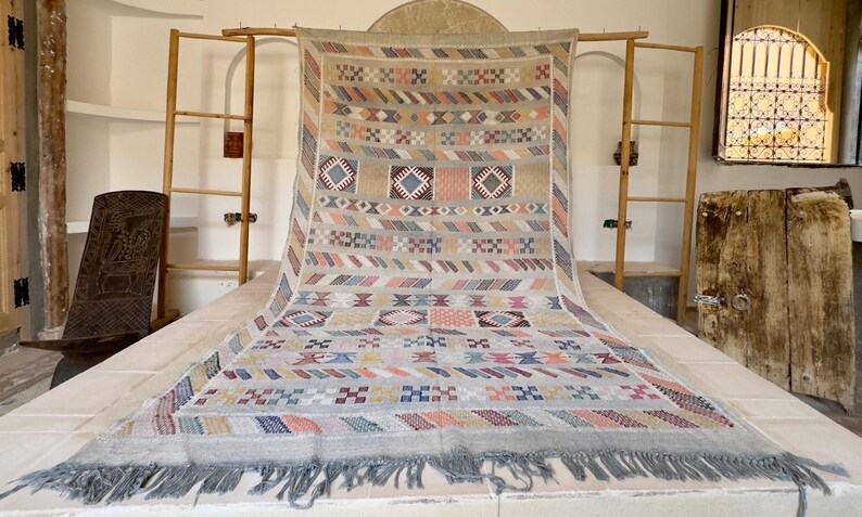May include: A large, rectangular rug with a beige background and colorful geometric patterns. The design includes squares, diamonds, and stripes in shades of blue, pink, orange, and green. The rug has a fringed edge.