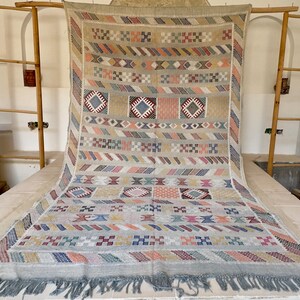 May include: A large, rectangular, handwoven rug with a geometric pattern in beige, blue, pink, orange, and gray. The rug has a fringe along the bottom edge and is displayed on a white surface.