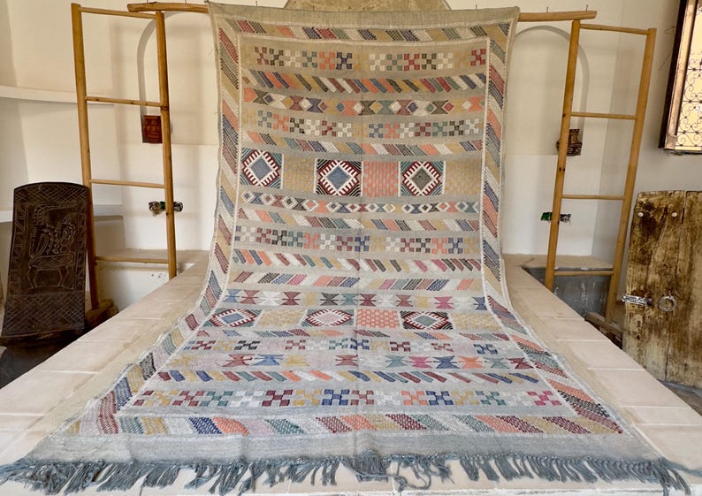 May include: A large, rectangular, handwoven rug with a geometric pattern in beige, blue, pink, orange, and gray. The rug has a fringe along the bottom edge and is displayed on a white surface.