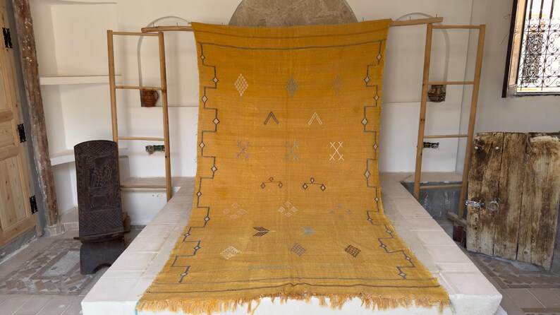 Pu&ograve; includere: Tappeto tessuto giallo senape con motivi geometrici e frange. Il tappeto presenta disegni a rombi, triangoli e croci in una tonalit&agrave; pi&ugrave; scura, con un bordo a zig-zag. Il tappeto &egrave; esposto in una stanza con scaffalature in legno e una porta rustica.