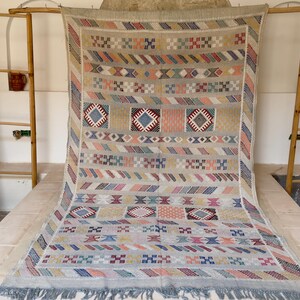 May include: A beige and multi-colored woven rug with geometric patterns. The rug features diamond, square, and stripe designs in shades of blue, pink, and beige. It has a fringed edge and is displayed on a light-colored surface.