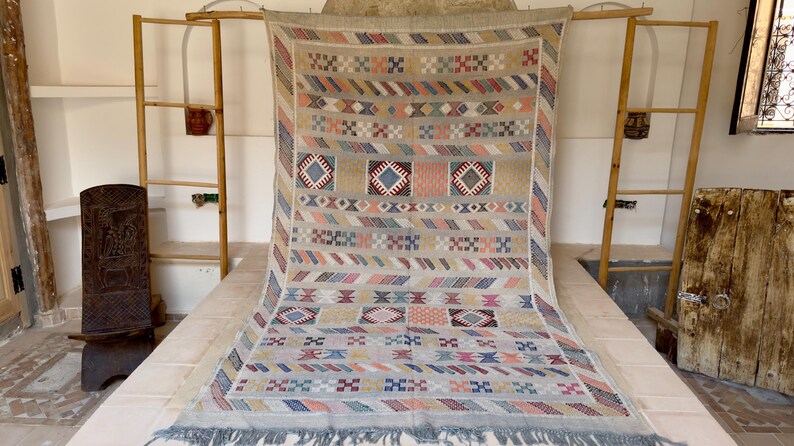 May include: A beige and multi-colored woven rug with geometric patterns. The rug features diamond, square, and stripe designs in shades of blue, pink, and beige. It has a fringed edge and is displayed on a light-colored surface.